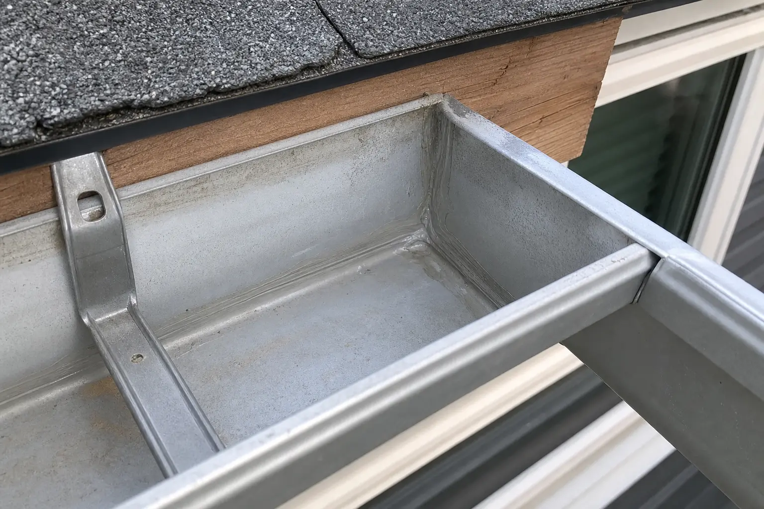 close-up of a residential gutter channel with visible joint sealant along an inside corner miter, aluminum surface showing light oxidation, wood fascia board visible behind gutter lip