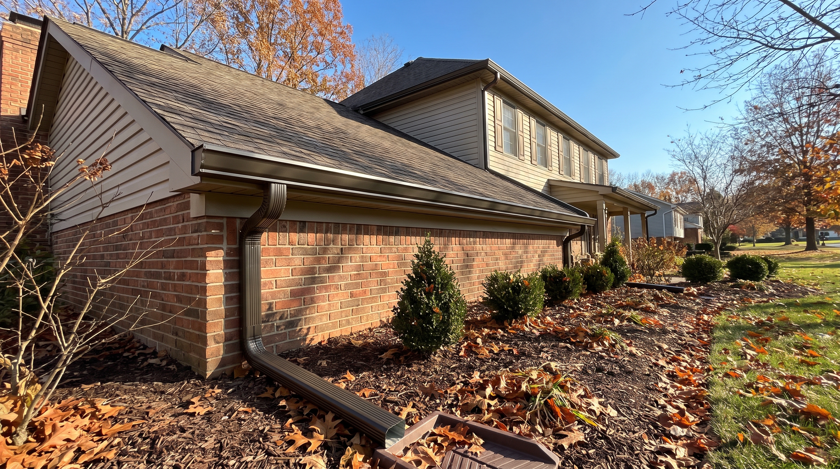 Comparison and Decision Guide — A residential home exterior in late autumn, showing a clean seamless aluminum gutter system freshly installed along the roofline with visible downspout extensions directing water away from a brick foundation, clear sky, organized landscaping (AI-generated)